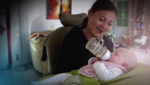 Jeune femme atteinte d'arthrogrypose, en fauteuil, donnant le biberon à son bébé